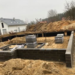 Fundamenty domu w trakcie budowy: częściowo wymurowane ściany z bloczków, zbrojenie, folia izolacyjna. W tle nowy dom i hałdy ziemi.