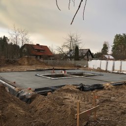 BUD-KOR Grzegorz Korgul - Zalany fundament z hydroizolacją i zbrojeniami na działce w Olsztynie. W tle domy jednorodzinne i drzewa. Widoczne materiały izolacyjne.