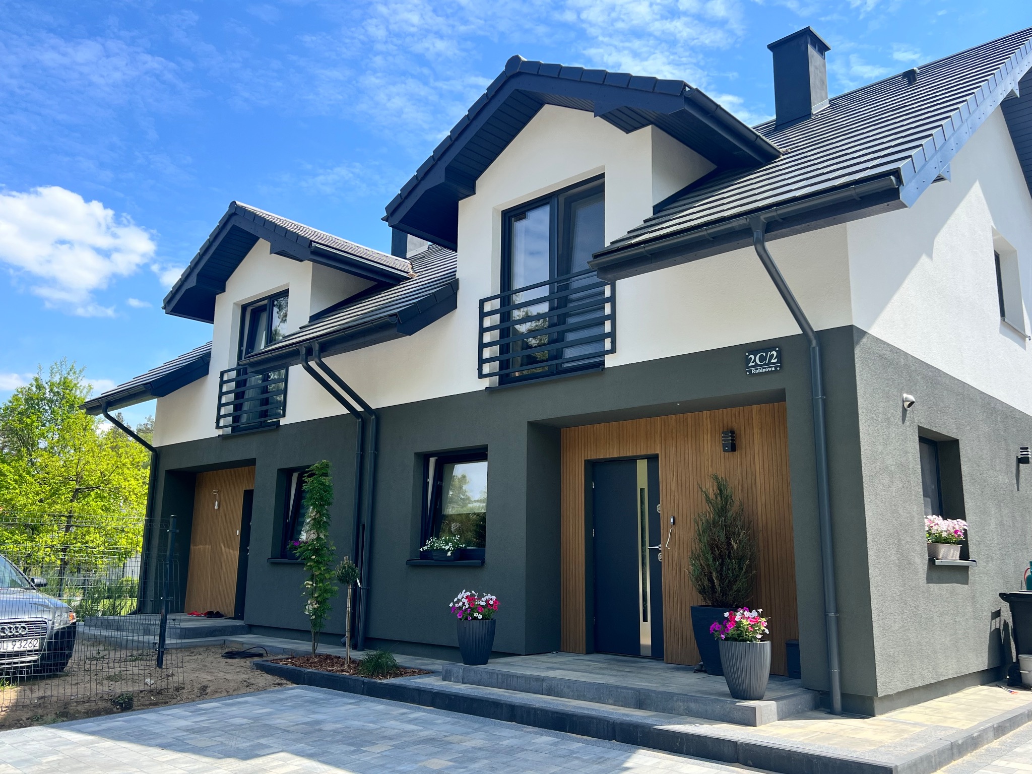 Nowoczesny bliźniak z ciemną elewacją i drewnianymi akcentami w Olsztynie. Widoczne balkony z czarną balustradą, zadbany teren wokół budynku i błękitne niebo.