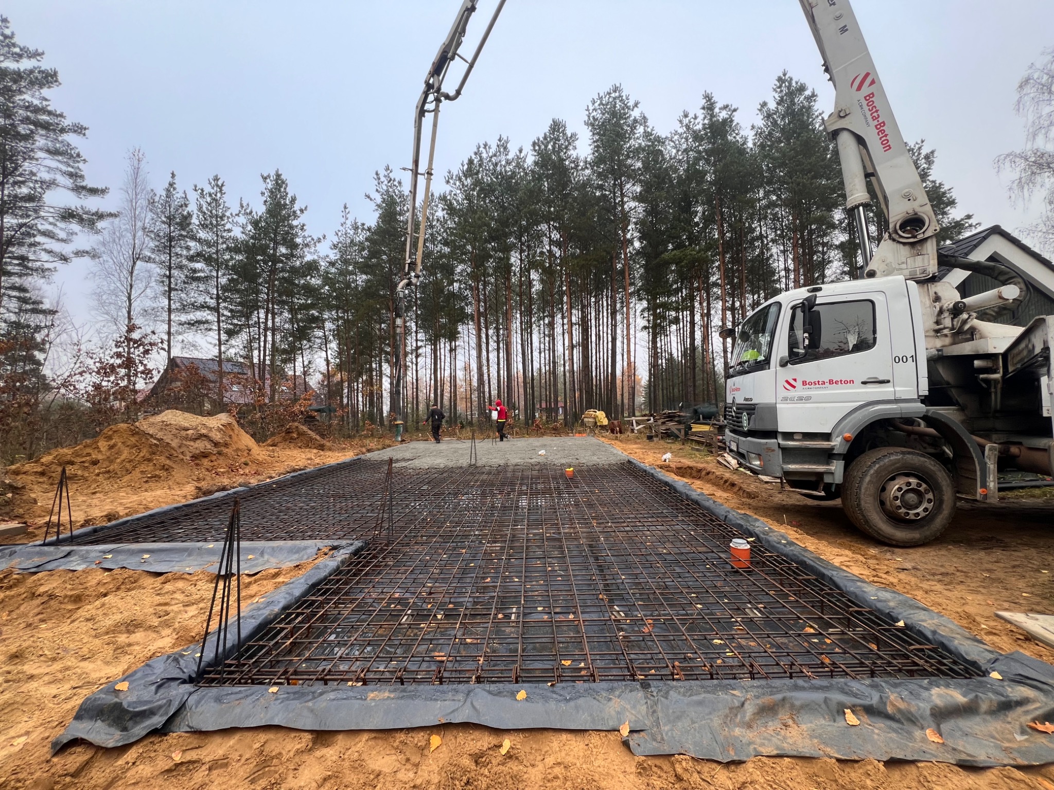 Zbrojenie fundamentu pod dom jednorodzinny, częściowo zalane betonem, z widocznym podajnikiem betonu Bosta-Beton i pracownikami w tle. Teren budowy na obrzeżach lasu.