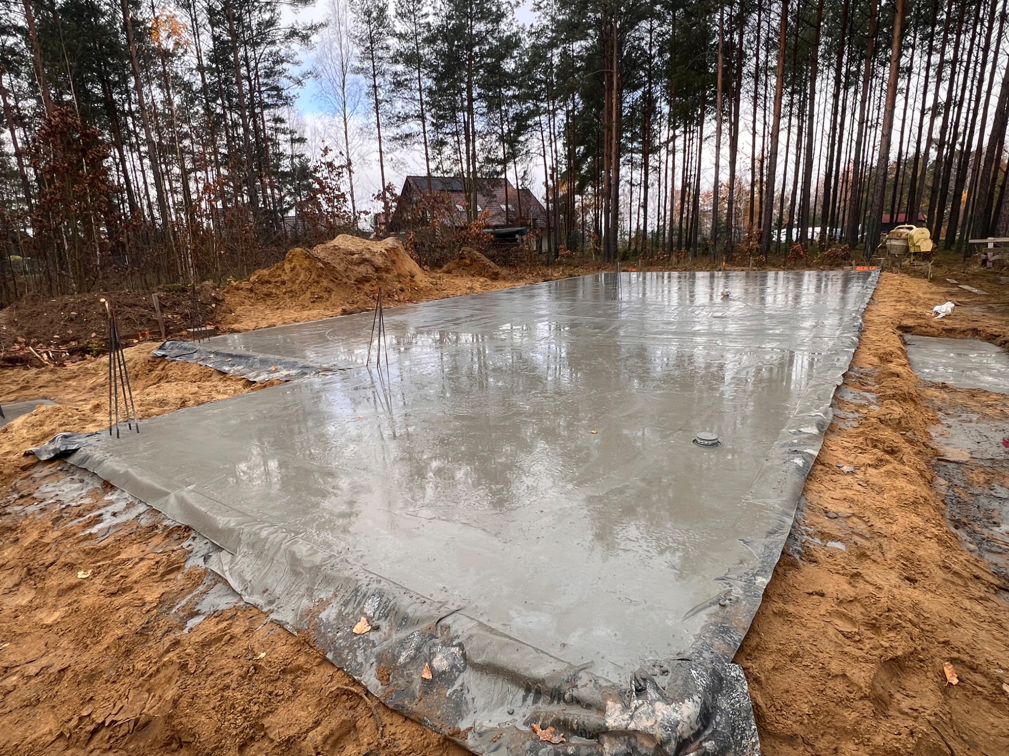 Wylana, mokra płyta fundamentowa na budowie domu jednorodzinnego, zabezpieczona folią. W tle las i częściowo widoczny budynek w stanie surowym.