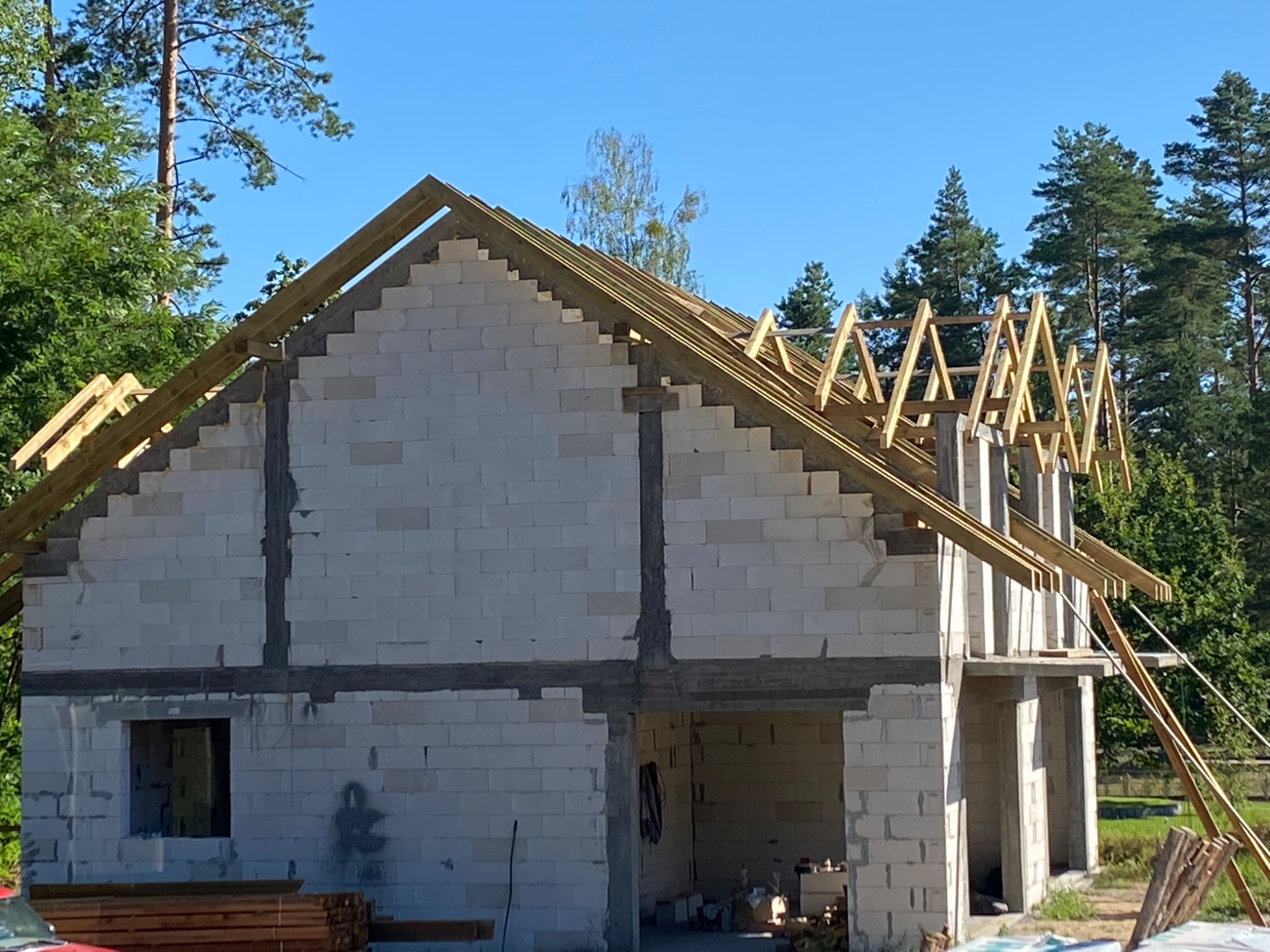 Wznoszenie dwukondygnacyjnego domu z białych bloczków, widoczne drewniane elementy konstrukcji dachu na tle błękitnego nieba i zieleni drzew.