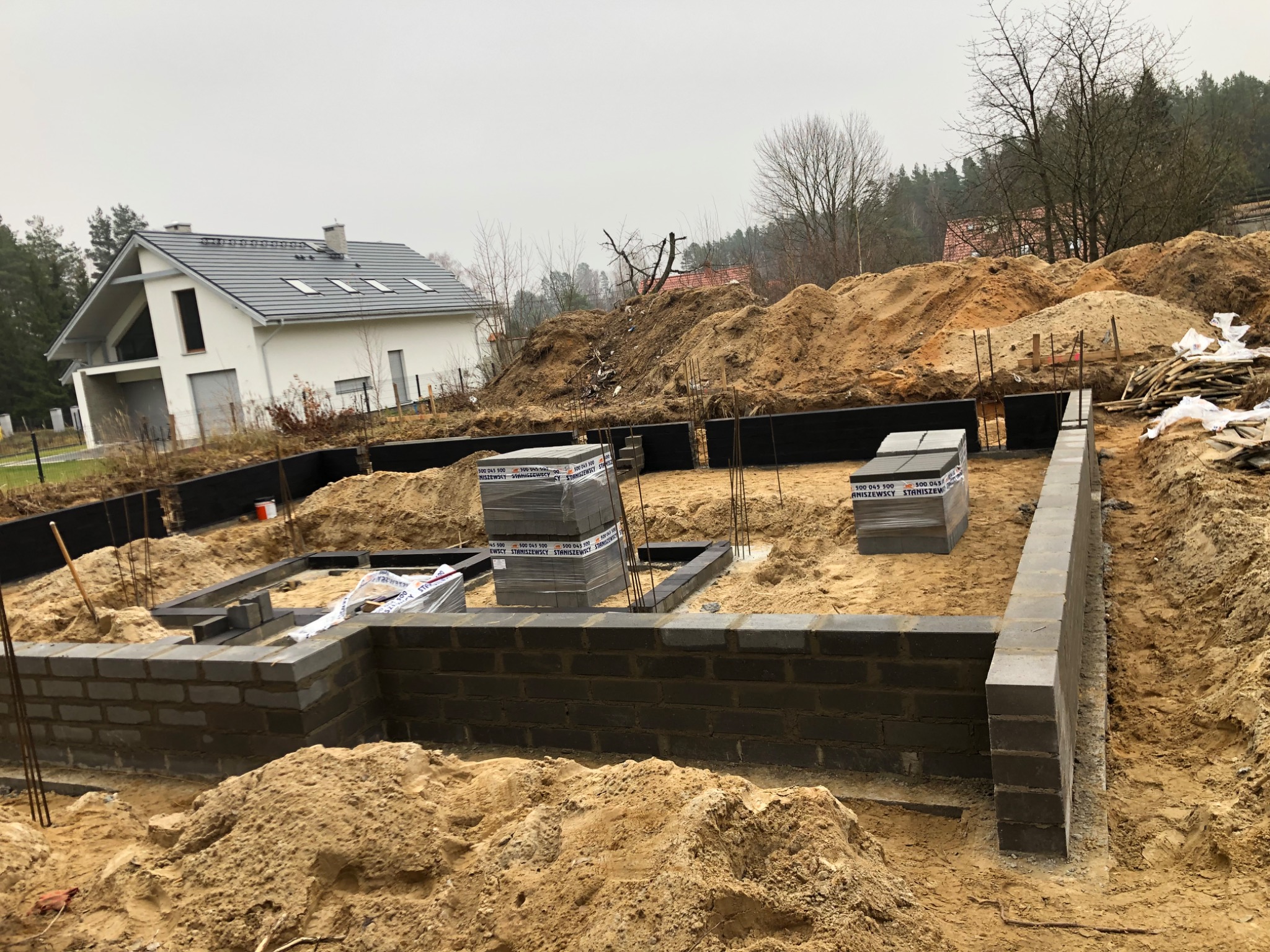 Fundamenty domu w trakcie budowy: częściowo wymurowane ściany z bloczków, zbrojenie, folia izolacyjna. W tle nowy dom i hałdy ziemi.