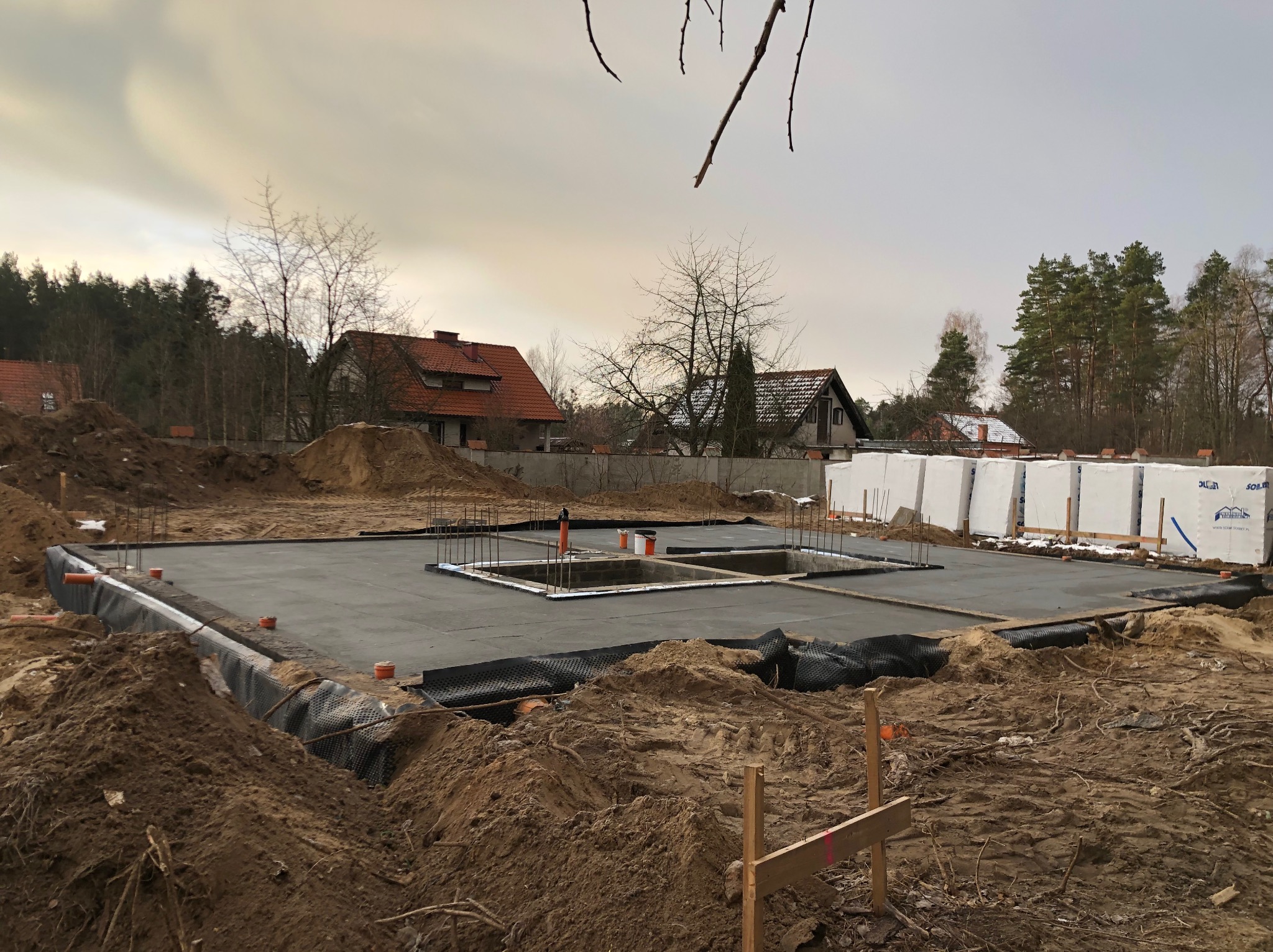 Zalany fundament z hydroizolacją i zbrojeniami na działce w Olsztynie. W tle domy jednorodzinne i drzewa. Widoczne materiały izolacyjne.