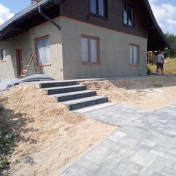K & E - BRUK BRUKARSTWO TRADYCYJNE I ARTYSTYCZNE EWA BUDNIK - Ułożona szara kostka brukowa prowadząca do schodów wejściowych domu z drewnianą elewacją. Widoczny piasek i prace wykończeniowe.