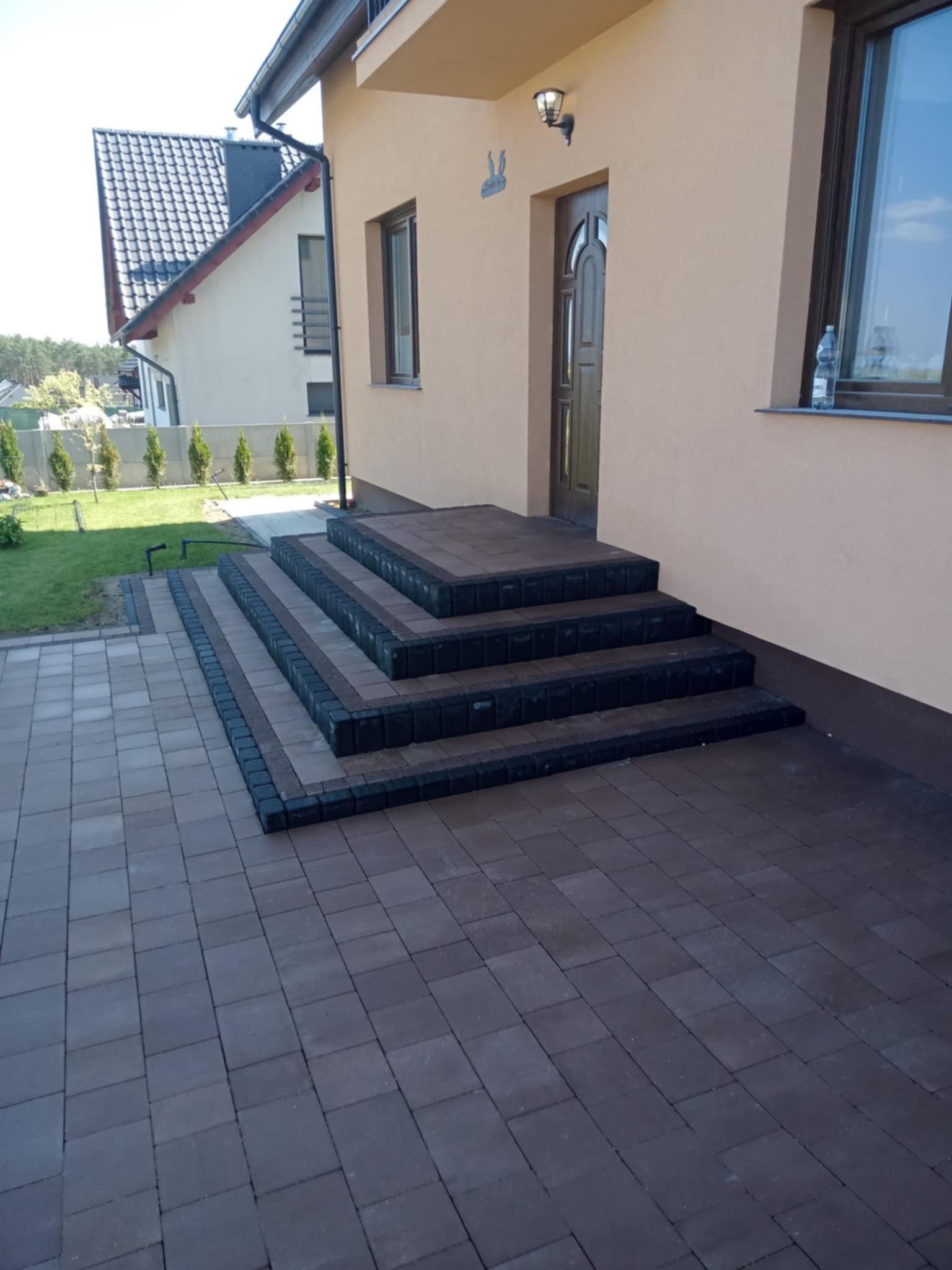Ułożona kostka brukowa w odcieniach brązu i szarości tworząca schody wejściowe do domu, z widocznym fragmentem chodnika i trawnika. Estetyczne wykończenie.