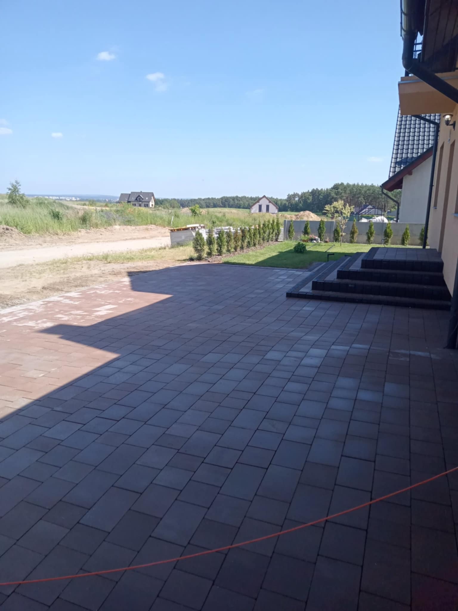 Ułożona, grafitowa kostka brukowa prowadząca do schodów wejściowych domu. W tle widok na okolicę i domy. Słoneczny dzień.