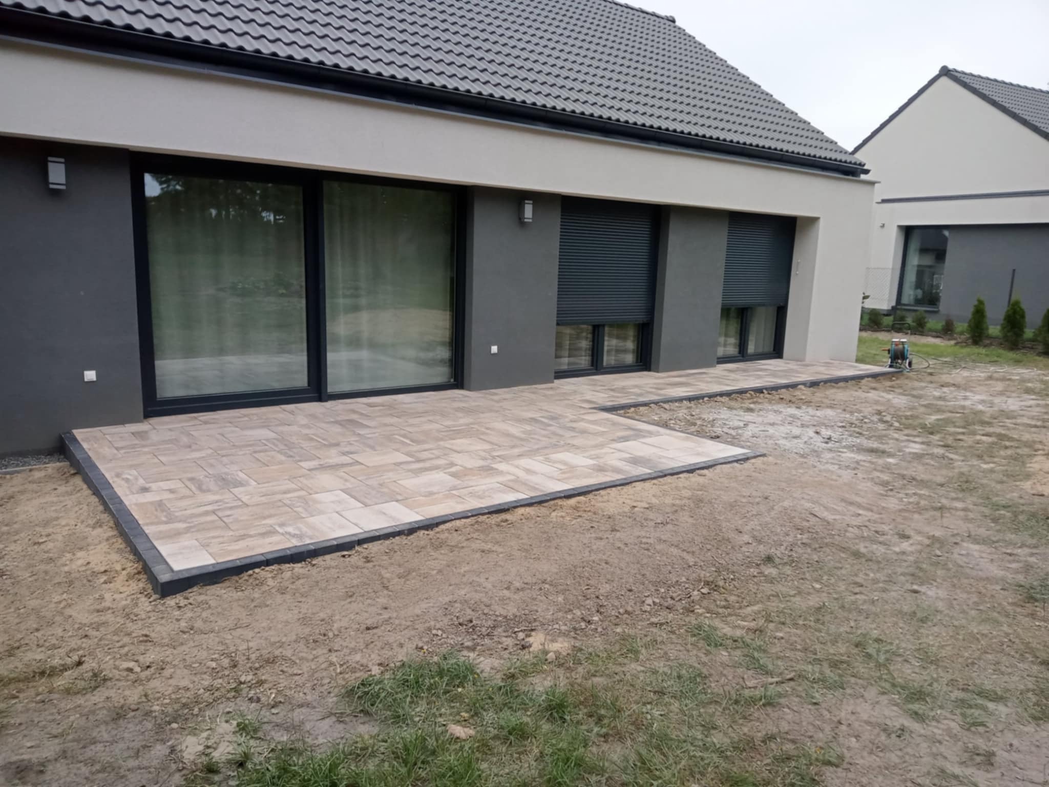Ułożona kostka brukowa na tarasie przylegającym do nowoczesnego domu z szarymi elementami elewacji i roletami zewnętrznymi. Widoczny fragment trawnika.