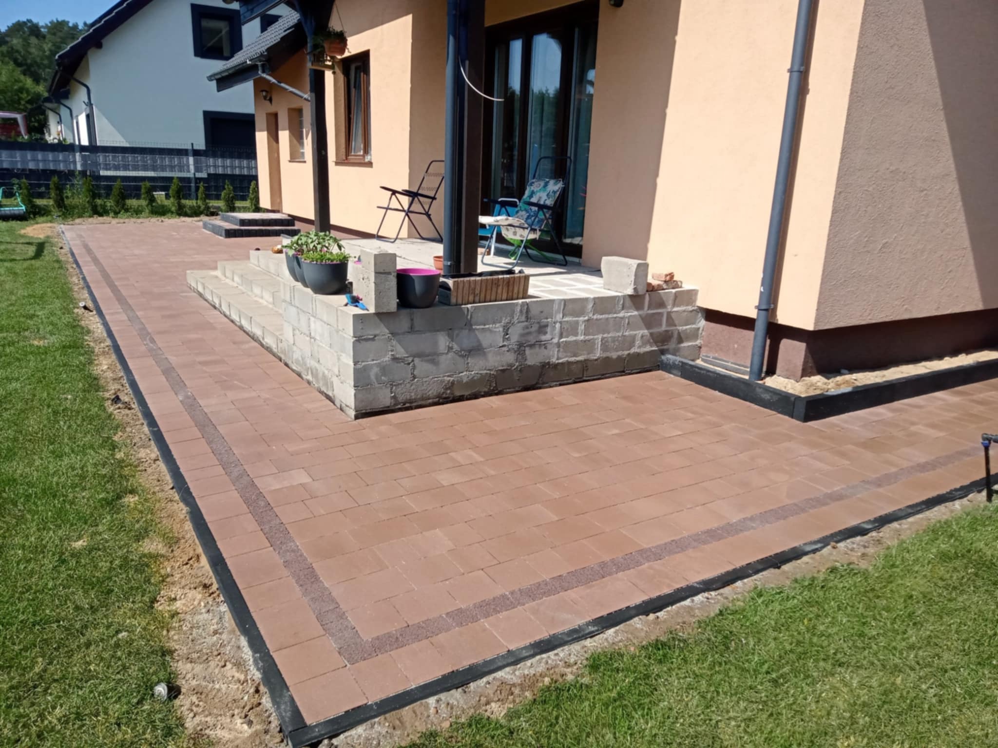 Wykończony taras z kostki brukowej w odcieniach brązu, z obrzeżami i schodami z bloczków betonowych. Widok na dom z ogrodem w słoneczny dzień.