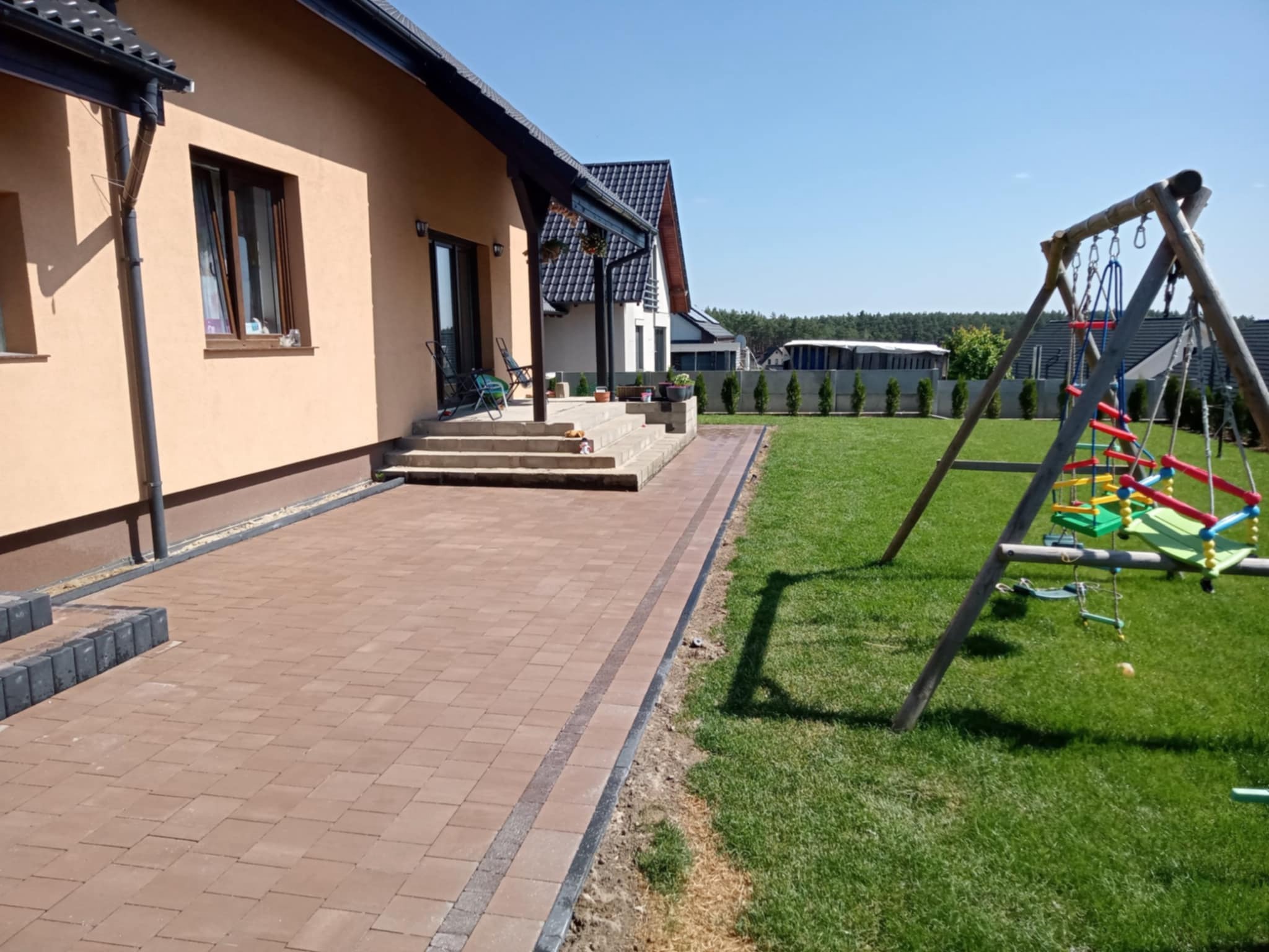 Brązowa kostka brukowa ułożona przy domu jednorodzinnym, obok trawnik z huśtawką. Widok na jasny, słoneczny dzień.