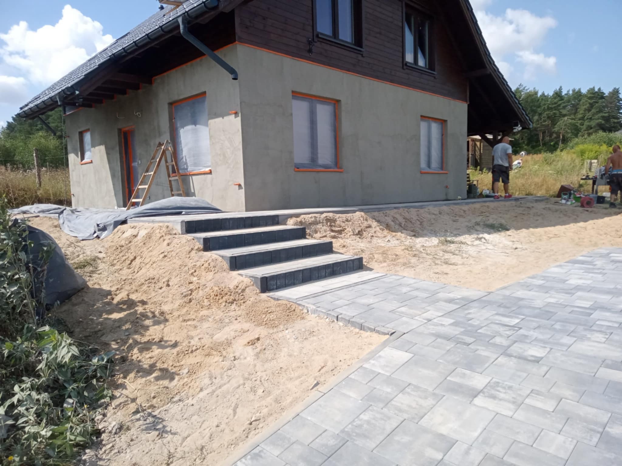 Ułożona szara kostka brukowa prowadząca do schodów wejściowych domu z drewnianą elewacją. Widoczny piasek i prace wykończeniowe.