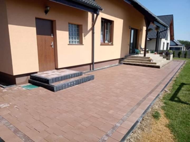 Brązowa kostka brukowa ułożona na podwórku domu jednorodzinnego, z obrzeżami i schodami z ciemnej kostki. Widok na elewację budynku i fragment ogrodu.