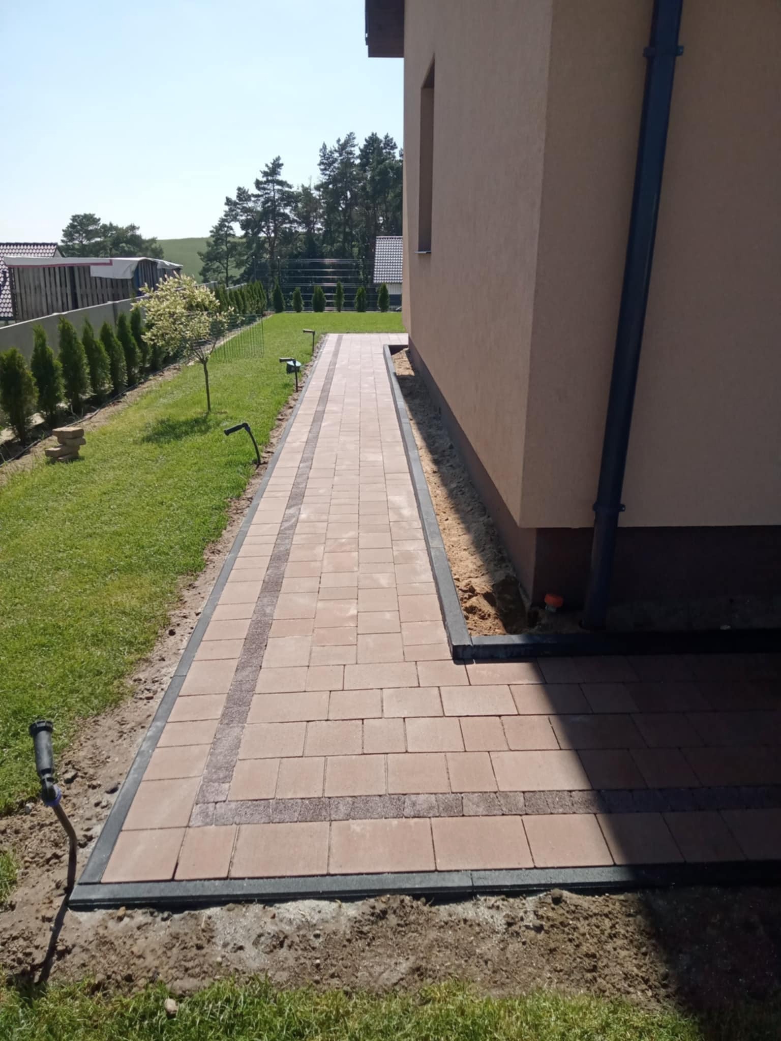 Ułożona ścieżka z kostki brukowej wzdłuż budynku, z widocznymi obrzeżami i trawnikiem z systemem nawadniania. Jasne, słoneczne ujęcie.