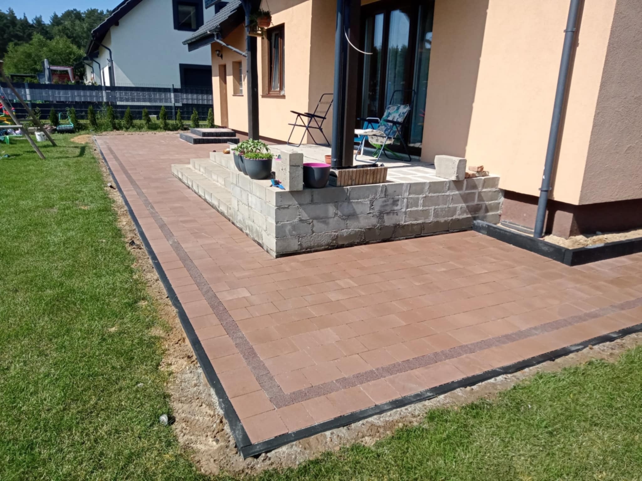 Wykończony taras z kostki brukowej w odcieniach brązu, z obrzeżami i schodami z bloczków betonowych, prowadzący do domu z jasną elewacją i zielonym trawnikiem.