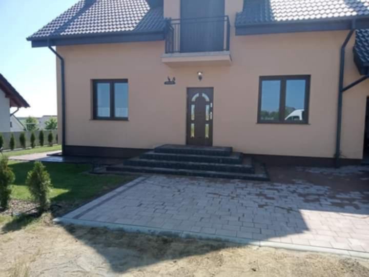 Ułożona kostka brukowa przed domem z beżową elewacją i schodami wejściowymi. Widoczne trawnik i tuje. Jasne, słoneczne oświetlenie.
