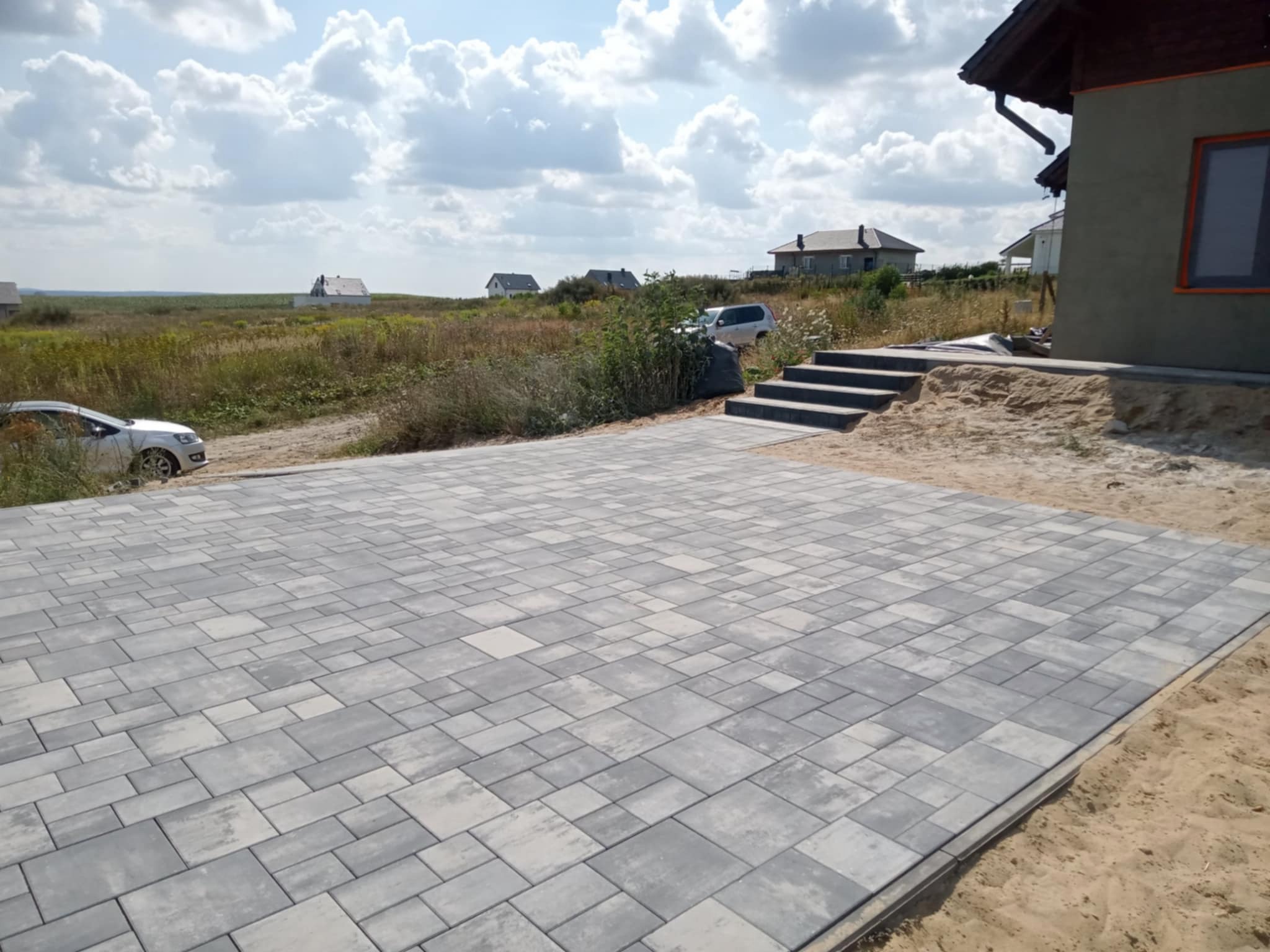 Ułożona szara kostka brukowa na podjeździe domu jednorodzinnego, widoczne schody wejściowe i trawnik z zabudową w tle, słoneczny dzień.