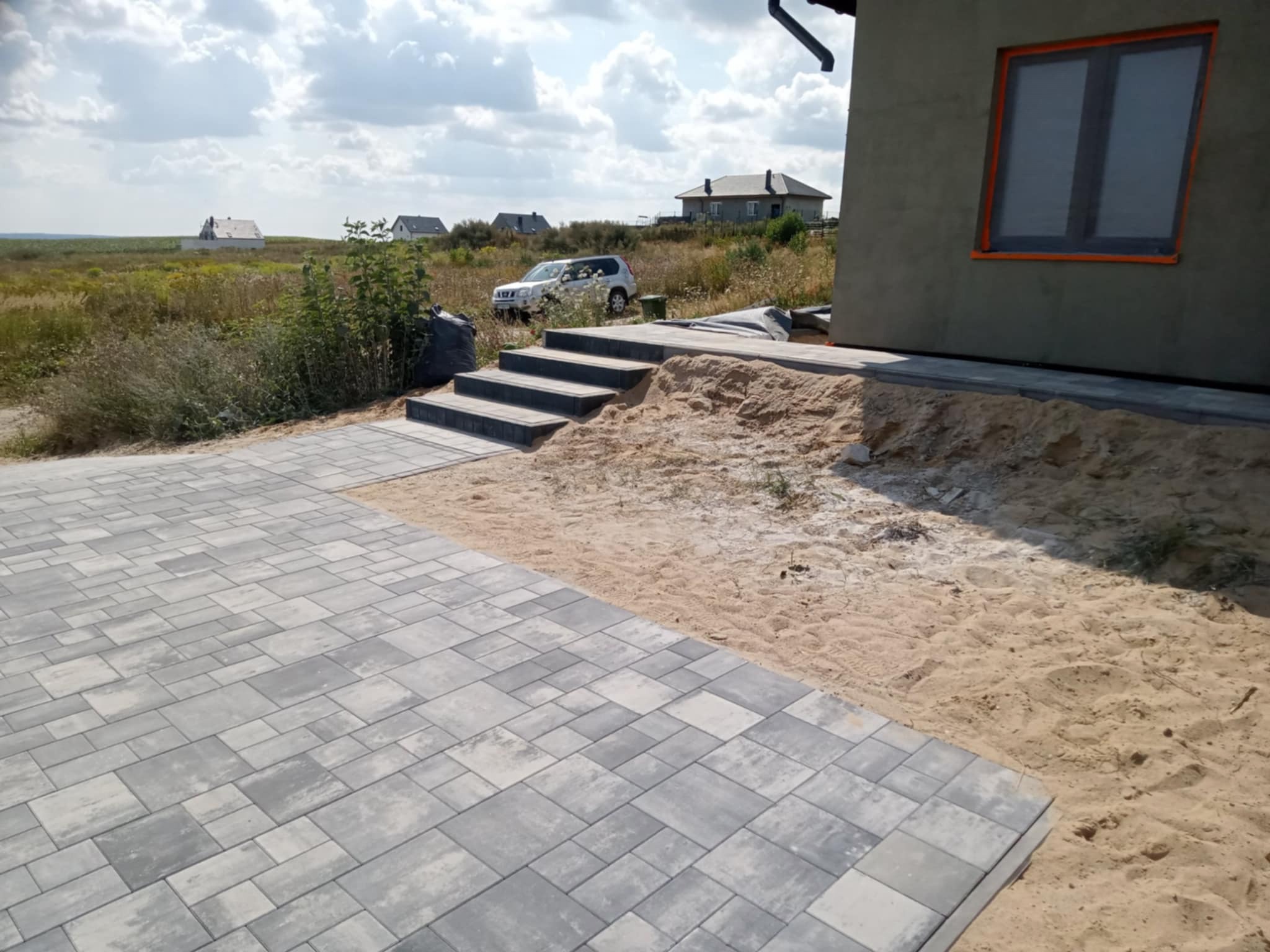Ułożona szara kostka brukowa przed domem w trakcie budowy, widoczne schody z kostki, piasek i samochód w tle. Słoneczny dzień, częściowe zachmurzenie.