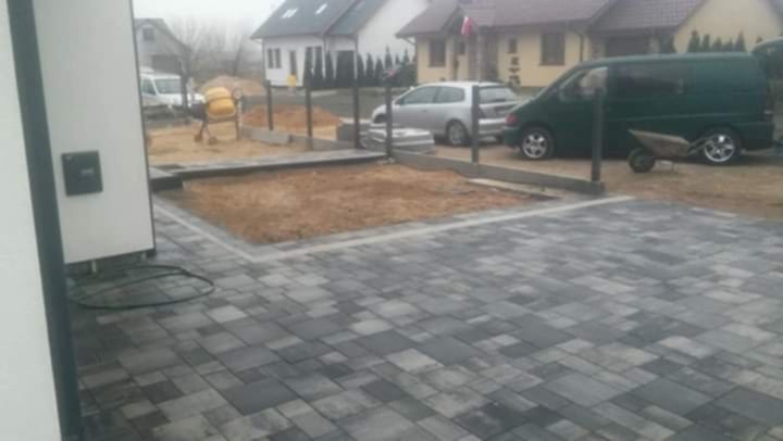 Ułożona kostka brukowa w odcieniach szarości, obrzeża z betonu, w tle teren przygotowany pod dalsze prace, betoniarka i samochody.