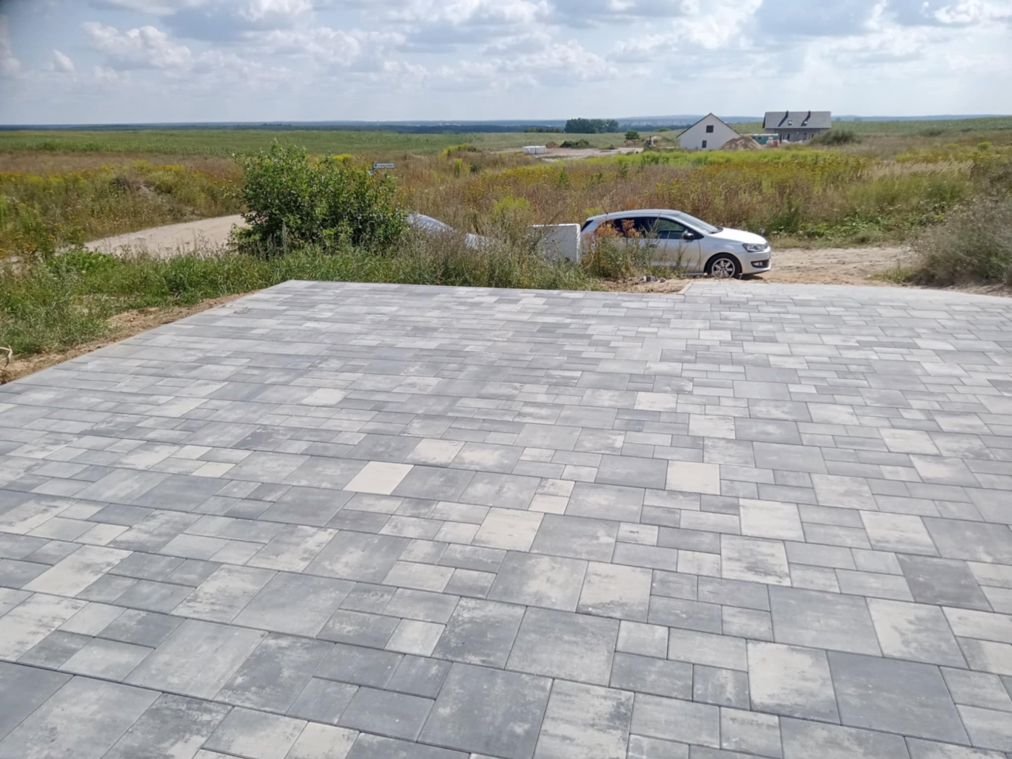 Ułożona kostka brukowa w odcieniach szarości na posesji, z widokiem na pole i domy w tle. Zaparkowany samochód osobowy. Słoneczny dzień.