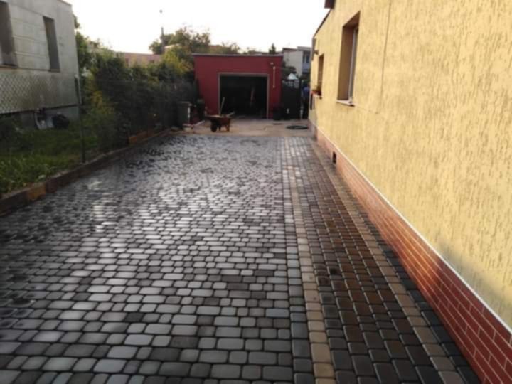 Ułożona kostka brukowa w odcieniach szarości i brązu tworzy wzór na podjeździe, przylegającym do budynku z żółtą elewacją i czerwoną podmurówką. W tle widoczny garaż.