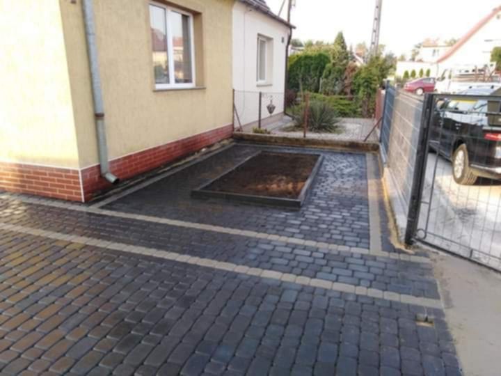 Ułożona, ciemnoszara kostka brukowa z jasnymi pasami, obok domu z ceglaną podmurówką i rabatą. Widoczna brama i fragment samochodu w tle.