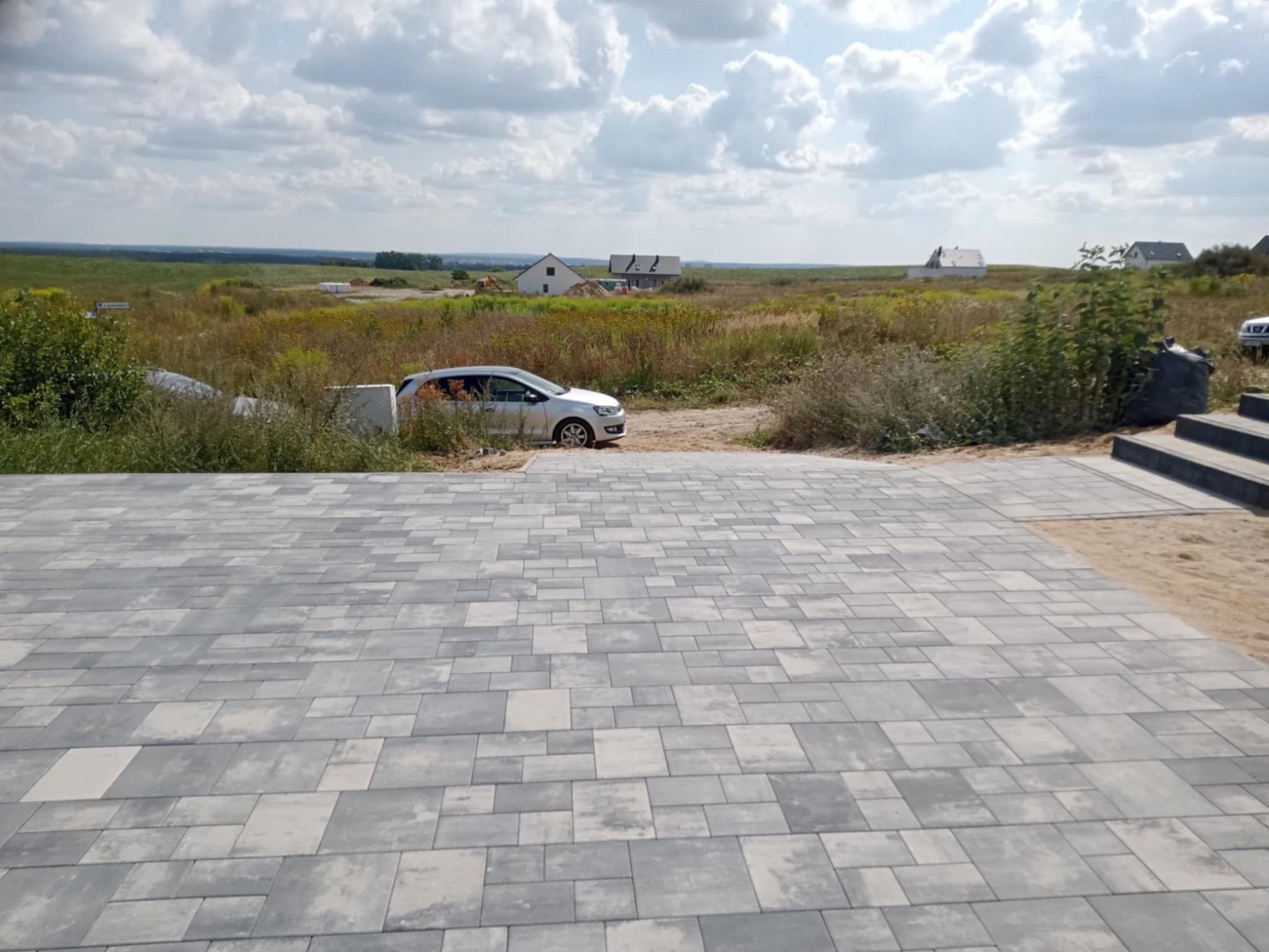 Ułożona kostka brukowa w odcieniach szarości, z widokiem na krajobraz i zabudowę w tle. Nowoczesny wzór kostki, jasne niebo z chmurami.