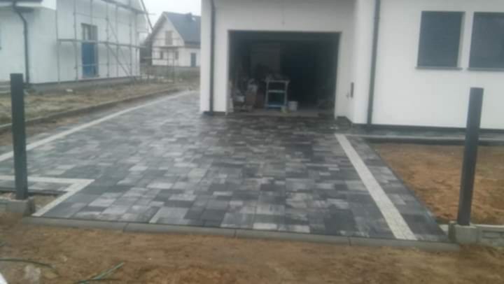 Ułożona, grafitowa kostka brukowa prowadząca do garażu, z jasnym pasem kostki po boku. Widoczne słupki ogrodzeniowe i fragment budynku w tle.