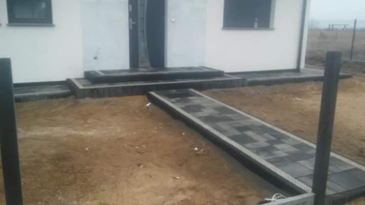 Ułożona ścieżka z kostki brukowej prowadząca do wejścia domu. Widoczne schody wejściowe również wyłożone kostką. Tło stanowi niezagospodarowany teren.