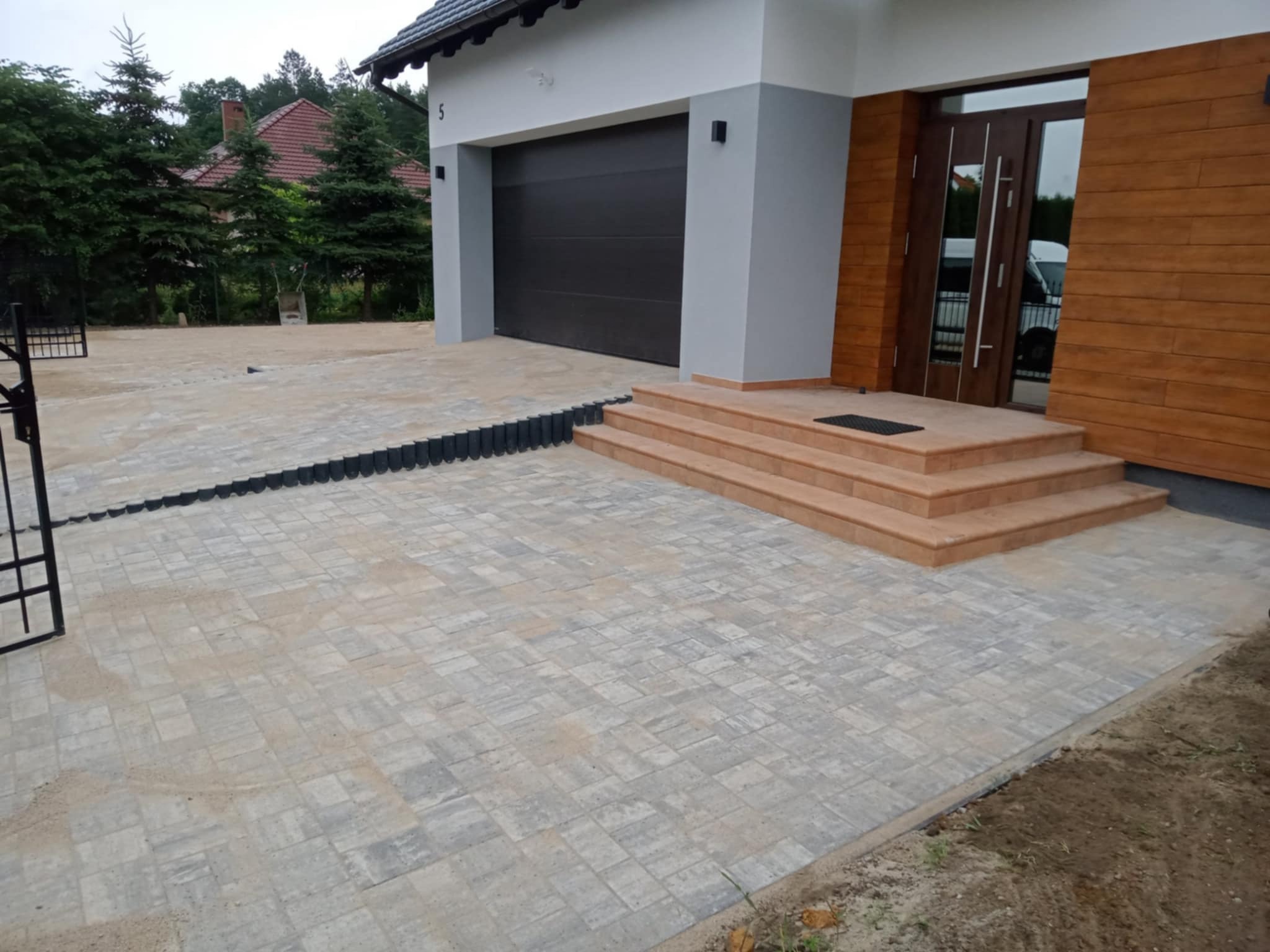 Ułożona kostka brukowa przed domem z garażem i schodami wejściowymi. Widoczne czarne obrzeża i fragment metalowej bramy. Tło stanowią drzewa i dom.