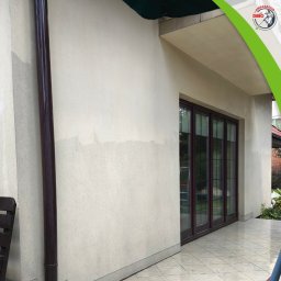 KRIS NA CIŚNIENIU Krzysztof Piwiński - Fragment elewacji domu z widoczną różnicą w czystości po umyciu, z brązową rynną i szklanymi drzwiami balkonowymi. Częściowo mokra, jasna kostka brukowa.