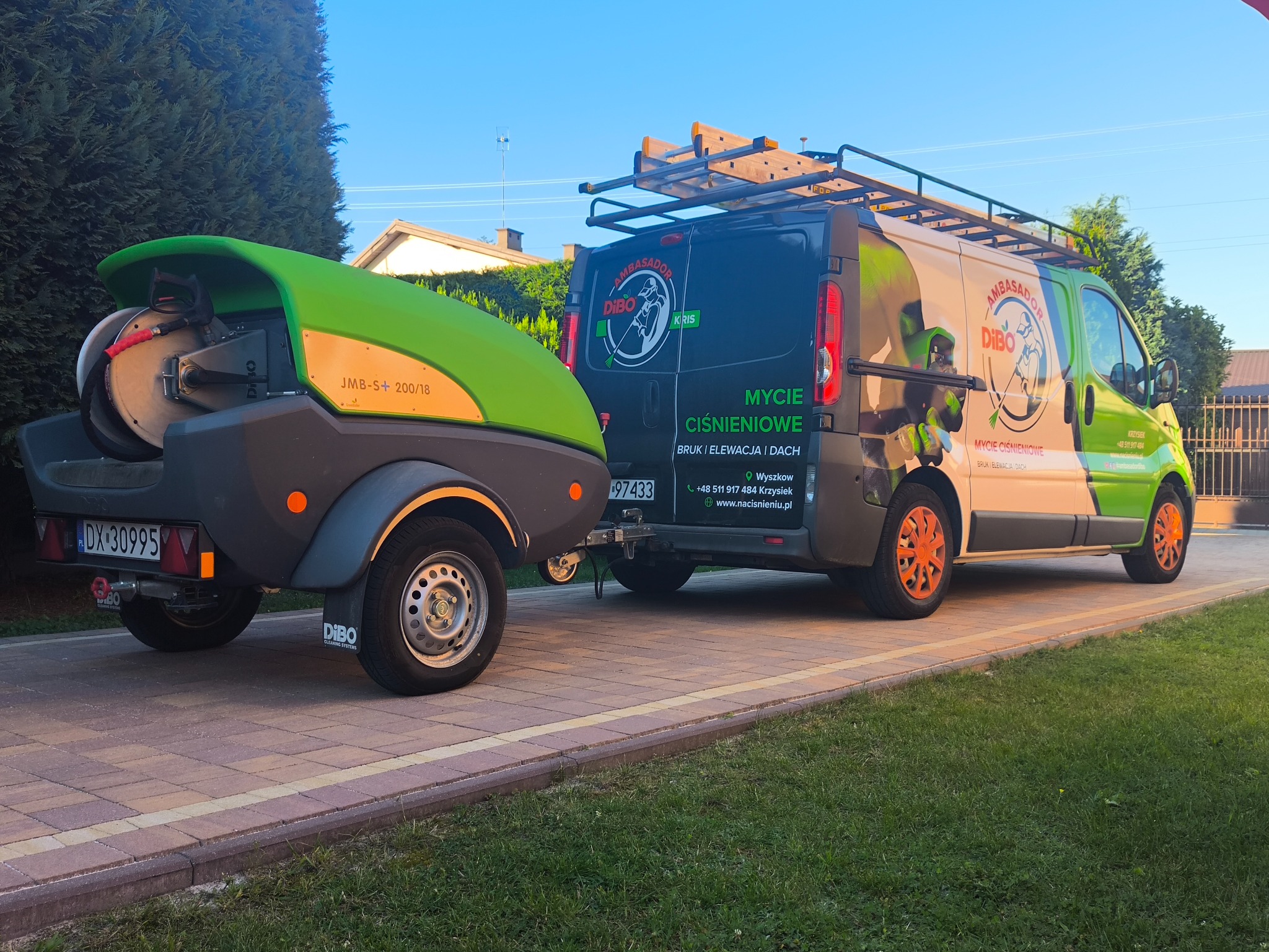 Przyczepa z myjką ciśnieniową Dibo podpięta do firmowego busa z reklamą mycia ciśnieniowego bruku, elewacji i dachów na tle zieleni i kostki brukowej.