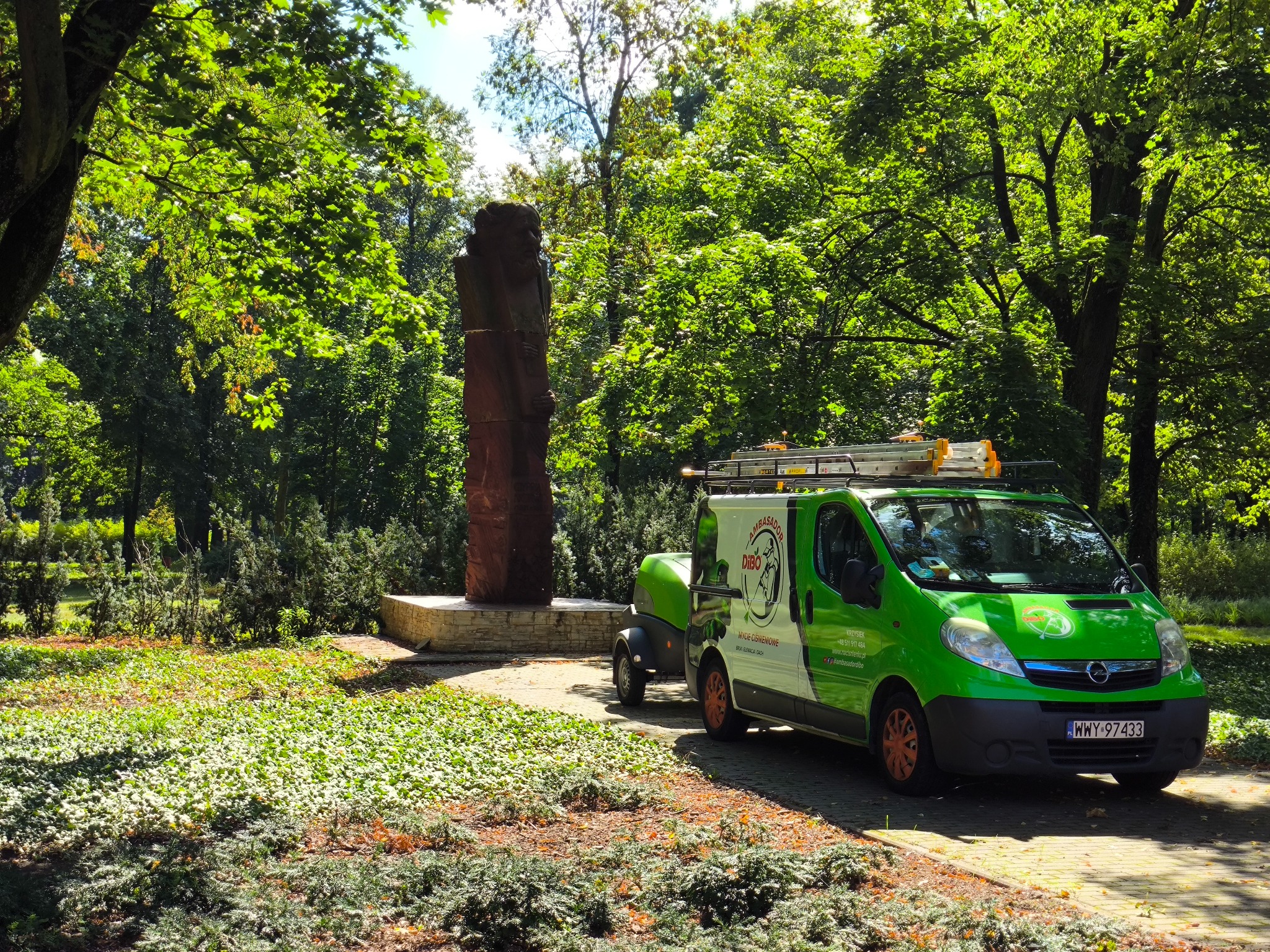 Zielony van z logo firmy czyszczącej DIBO, zaparkowany w parku obok pomnika. Na dachu drabina. Widoczne dane kontaktowe i hasło: Mycie ciśnieniowe.