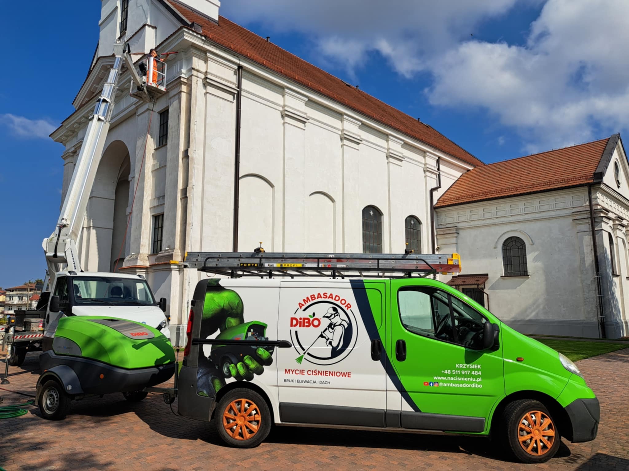 Pracownik na podnośniku czyści elewację kościoła. Na pierwszym planie pojazd z logo firmy oferującej mycie ciśnieniowe. Błękitne niebo w tle.