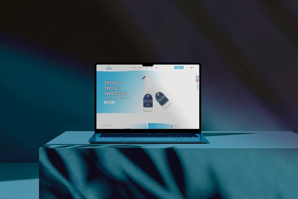 Laptop prezentuje stronę internetową marki Joanna z hasłem 'Źródło Twojej Świeżości' na tle ciemnego, oświetlonego studia z cieniem liścia palmy. Minimalistyczny design.