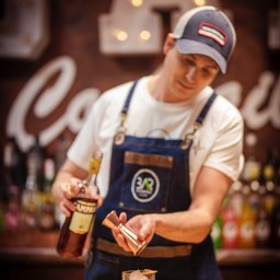 BarCocktail - Barman w denimowym fartuchu przygotowuje drinka Campari z lodem i skórką pomarańczy, używając miarki barmańskiej. W tle butelki i dekoracja z żarówkami.