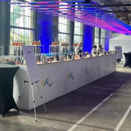 BarCocktail - Długa, biała lada barowa z menu na statywie, za nią barmani przygotowują drinki. W tle industrialne okna i dekoracyjne oświetlenie. Event firmowy w hali.