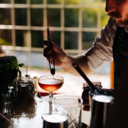 BarCocktail - Barman w zielonym fartuchu przygotowuje koktajl, używając szczypiec do dodania wisienki do kieliszka. W tle okno z widokiem na zieleń.