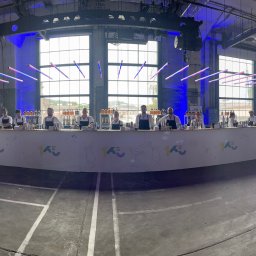 BarCocktail - Szeroki kadr na bar z obsługą podczas eventu firmowego. W tle industrialne wnętrze z dużymi oknami i dekoracyjnym oświetleniem. Na barze widoczne butelki i akcesoria barmańskie.