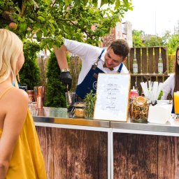 BarCocktail - Mobilny bar na zewnątrz: barman w akcji przygotowuje drinka dla klientki, obok stoi barmanka z gotowymi koktajlami. Dekoracje z drewna i zieleni tworzą naturalny klimat.