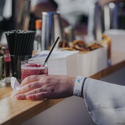 BarCocktail - Bar z koktajlem malinowym, słomką i dekoracją. W tle akcesoria barmańskie, serwetki i przekąski. Na pierwszym planie dłoń z opaską eventową.