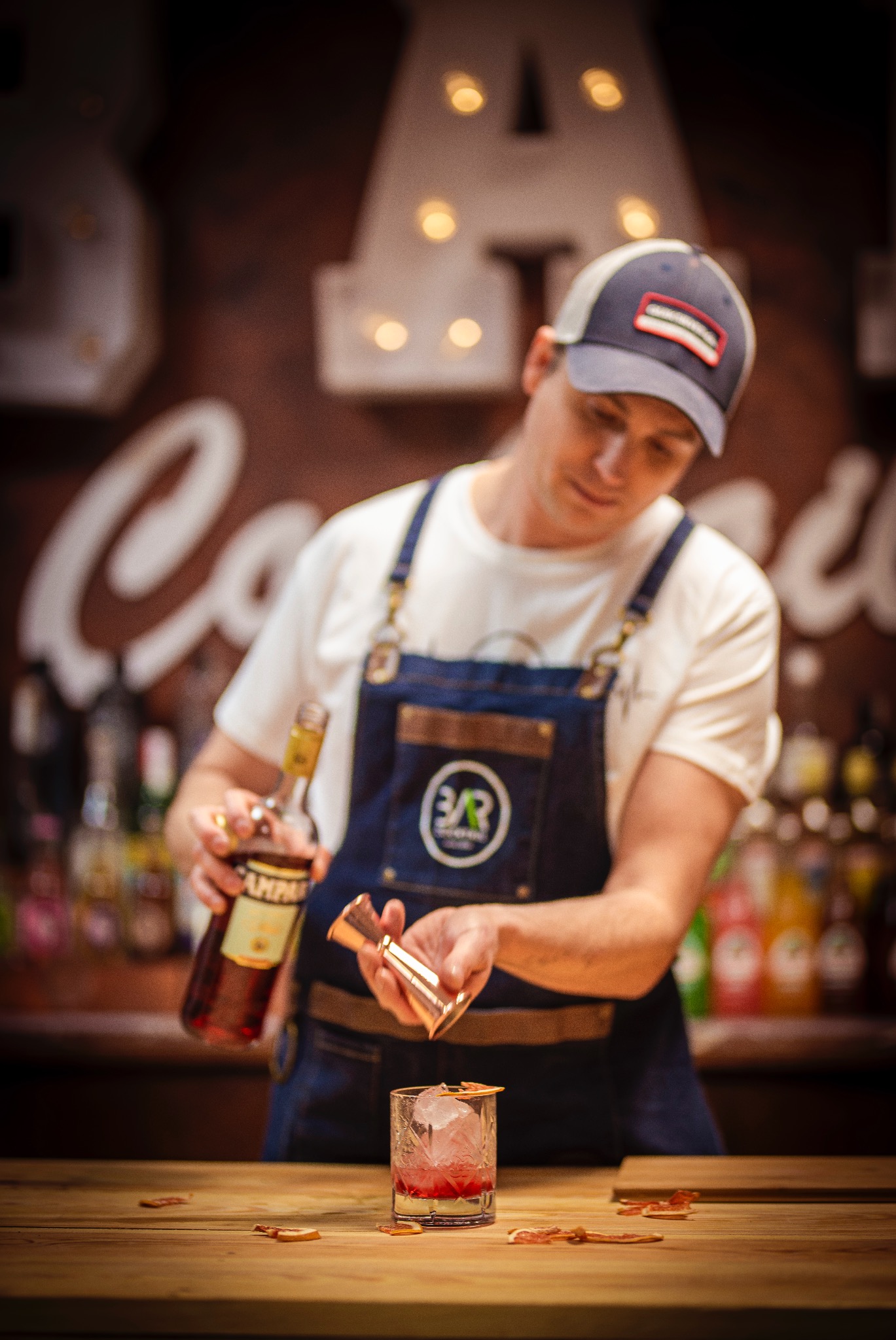 Barman w denimowym fartuchu przygotowuje drinka Campari z lodem i skórką pomarańczy, używając miarki barmańskiej. W tle butelki i dekoracja z żarówkami.