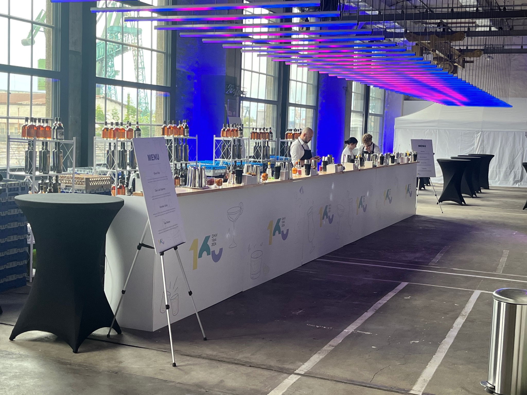 Długa, biała lada barowa z menu na statywie, za nią barmani przygotowują drinki. W tle industrialne okna i dekoracyjne oświetlenie. Event firmowy w hali.
