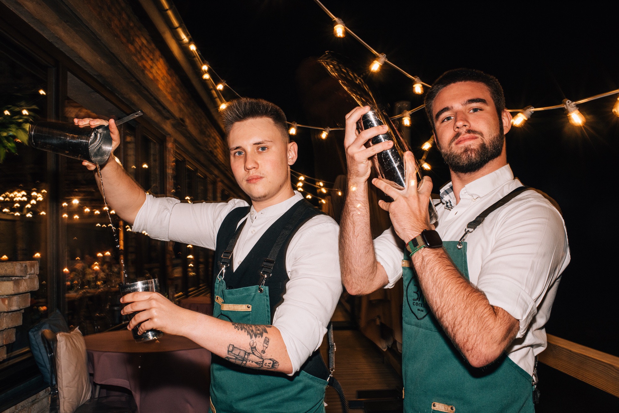 Dwóch barmanów w akcji, jeden przelewa drink z shakera do szklanki, drugi potrząsa shakerem, w tle dekoracyjne lampki. Profesjonalna obsługa barmańska na evencie.
