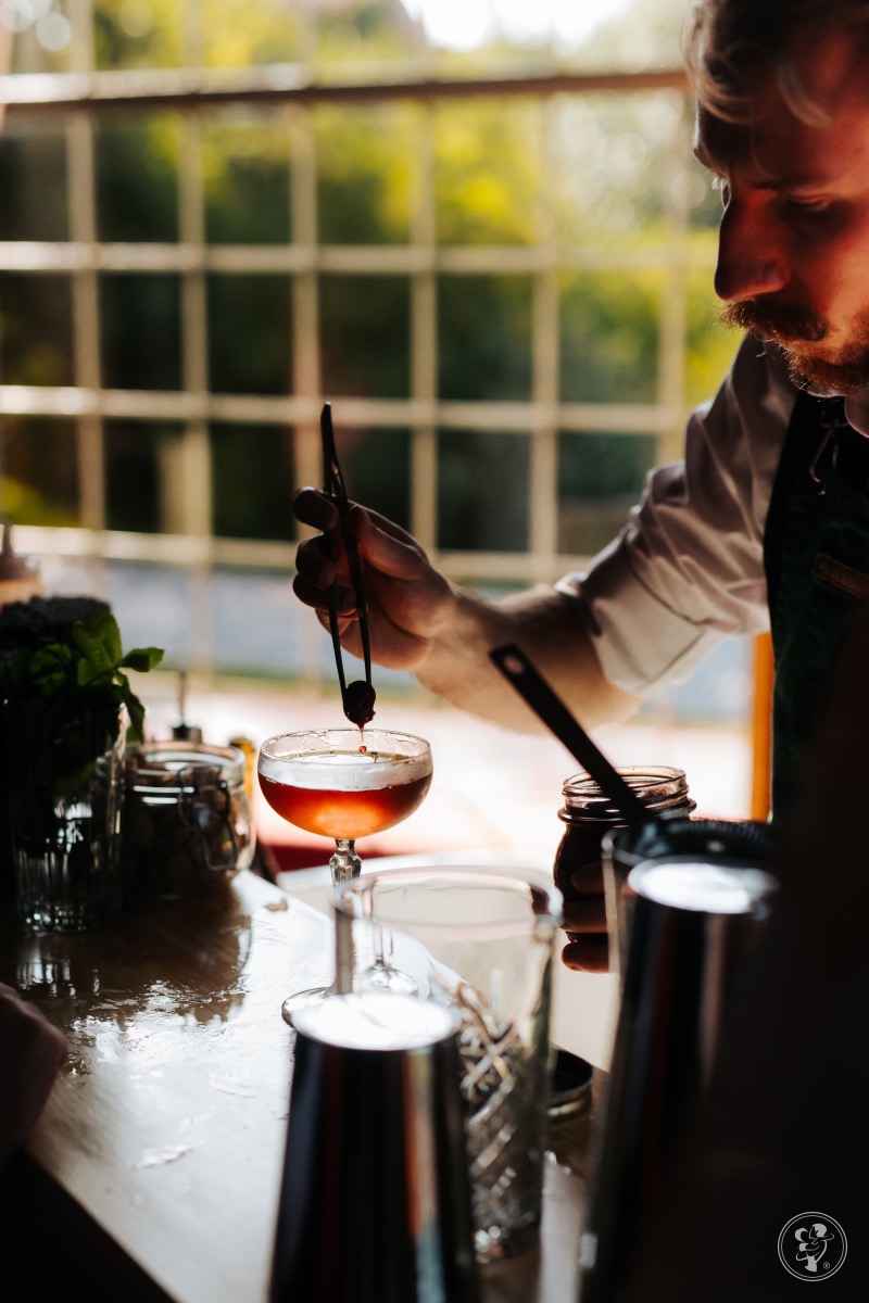 Barman w zielonym fartuchu przygotowuje koktajl, używając szczypiec do dodania wisienki do kieliszka. W tle okno z widokiem na zieleń.