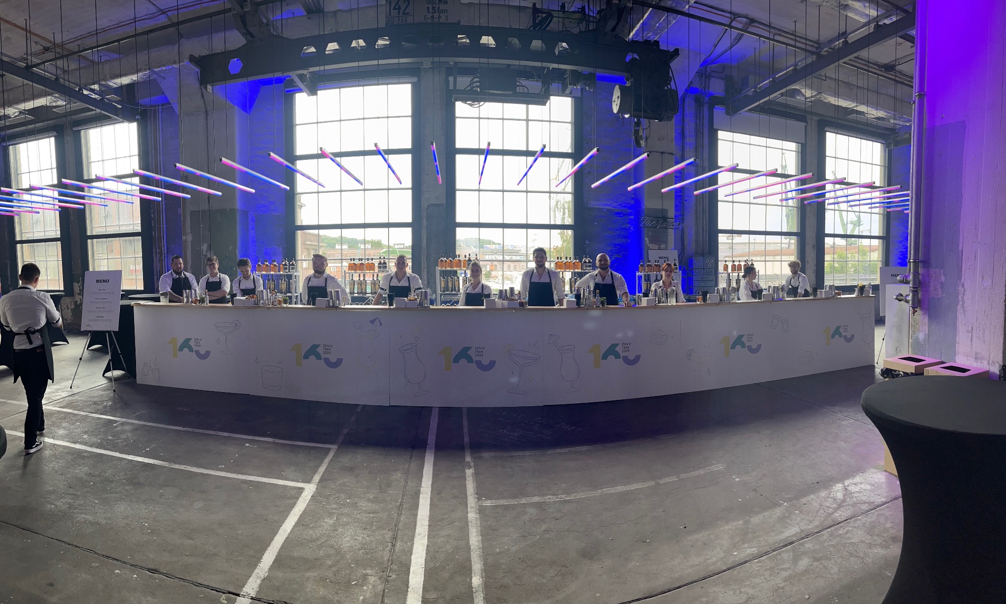 Szeroki kadr na bar z obsługą podczas eventu firmowego. W tle industrialne wnętrze z dużymi oknami i dekoracyjnym oświetleniem. Na barze widoczne butelki i akcesoria barmańskie.