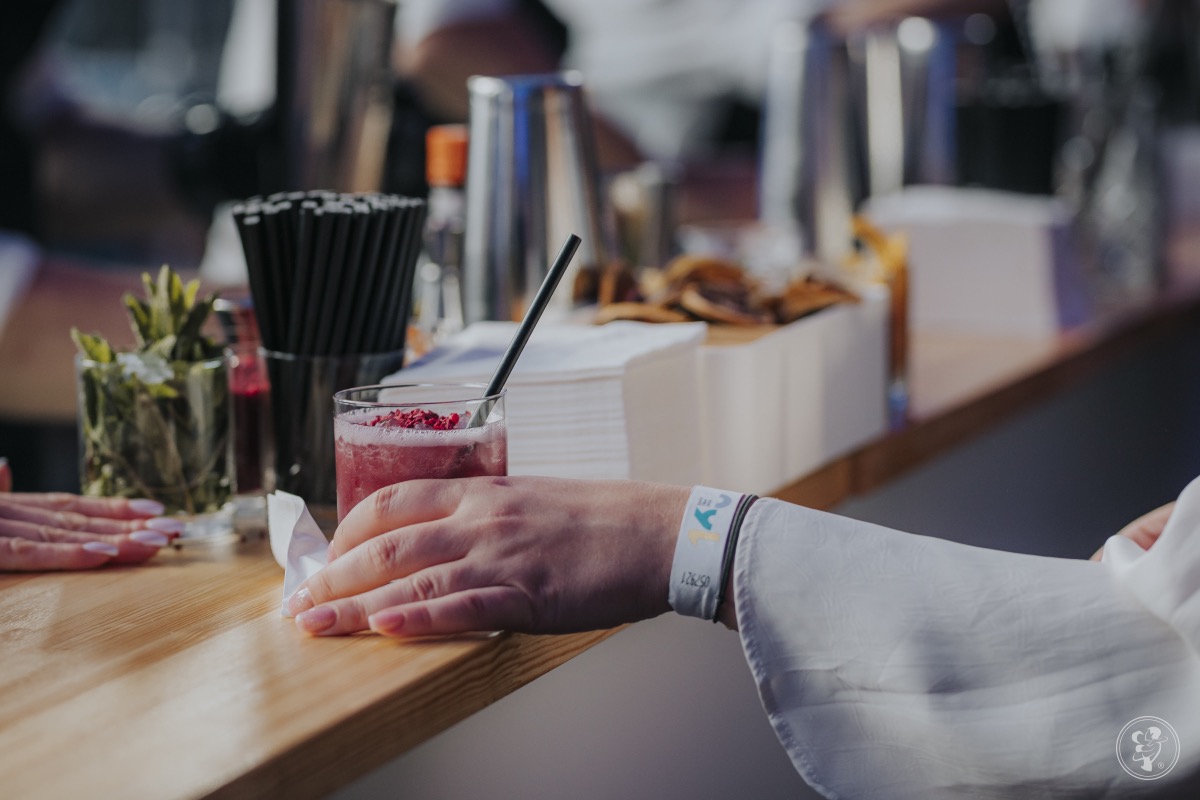 Bar z koktajlem malinowym, słomką i dekoracją. W tle akcesoria barmańskie, serwetki i przekąski. Na pierwszym planie dłoń z opaską eventową.