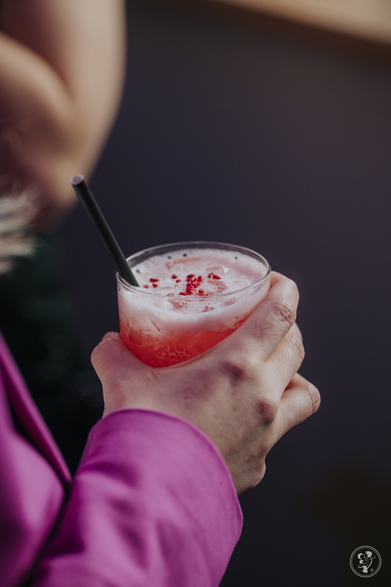 Różowy koktajl z czerwoną posypką, trzymany w dłoni kobiety w fuksjowej marynarce, z czarną słomką. Elegancki drink na przyjęcie.
