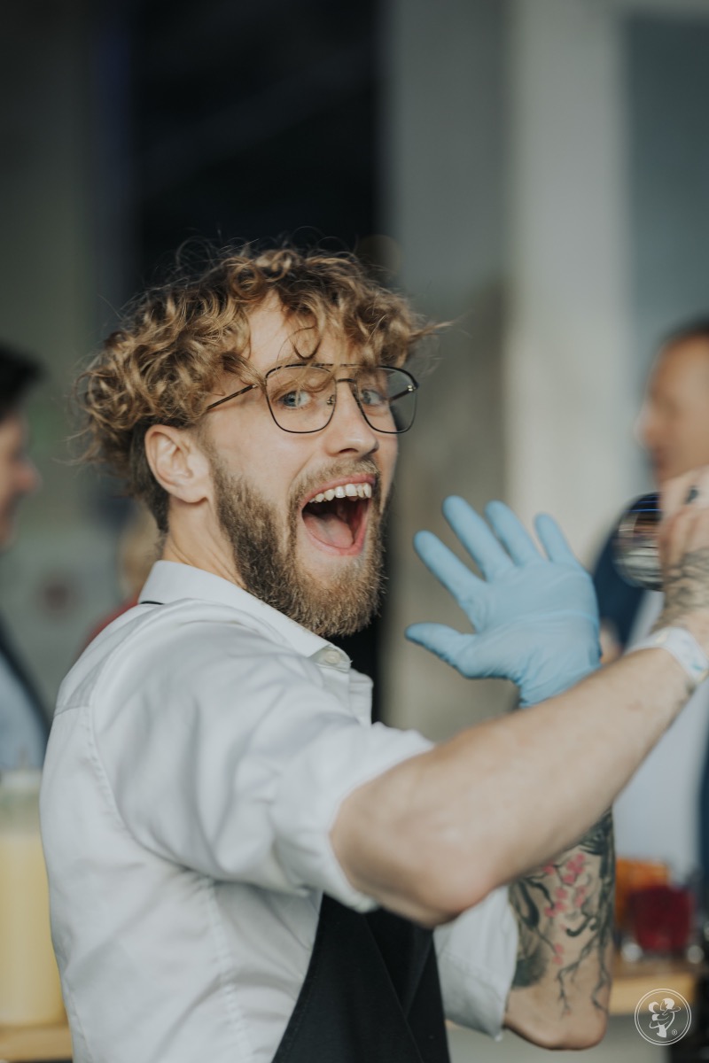Uśmiechnięty barman w okularach i rękawiczkach, z tatuażem na ramieniu, przygotowuje drinka na imprezie. W tle widoczni goście.