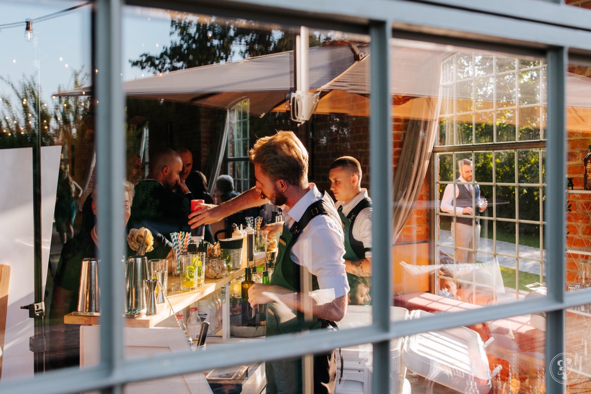 Barman w zielonym fartuchu przygotowuje drinki na przyjęciu plenerowym, widoczne szkło barowe i goście w tle, ujęcie przez okno z refleksami.