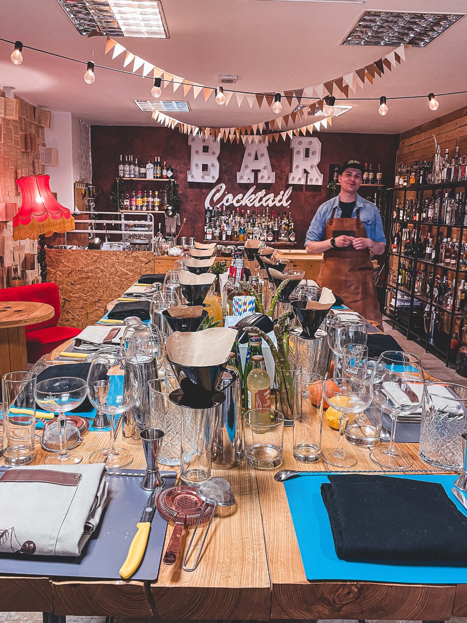 Przygotowany stół do warsztatów barmańskich: szkło, narzędzia, dekoracje świetlne i barman w tle. Klimatyczne oświetlenie i rustykalny wystrój.